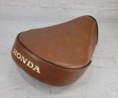 A brown Super Cub seat cover with gold 'HONDA' lettering, shown on a white wooden surface against a white brick wall background.