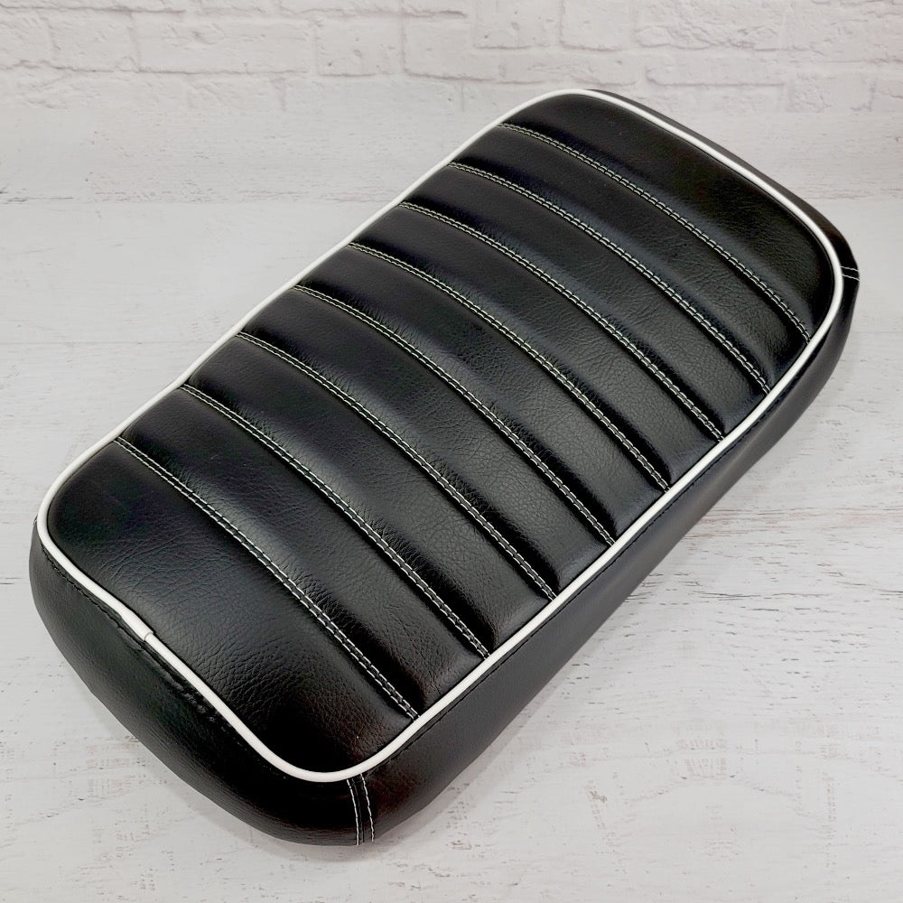 Black tuck-and-roll padded Maddog seat cover with horizontal pleats and white piping against a white brick backdrop, handmade by Cheeky Seats